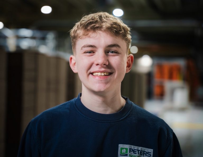 Ein junger, lachender Auszubildender von #TEAMFritzPeters steht in der Produktionshalle in Moers.